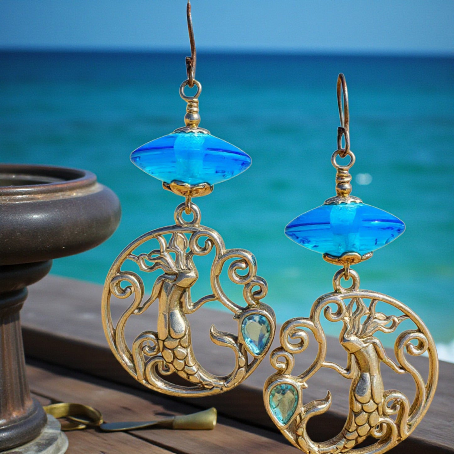 Mermaid-themed earrings with blue gemstones against a beach background