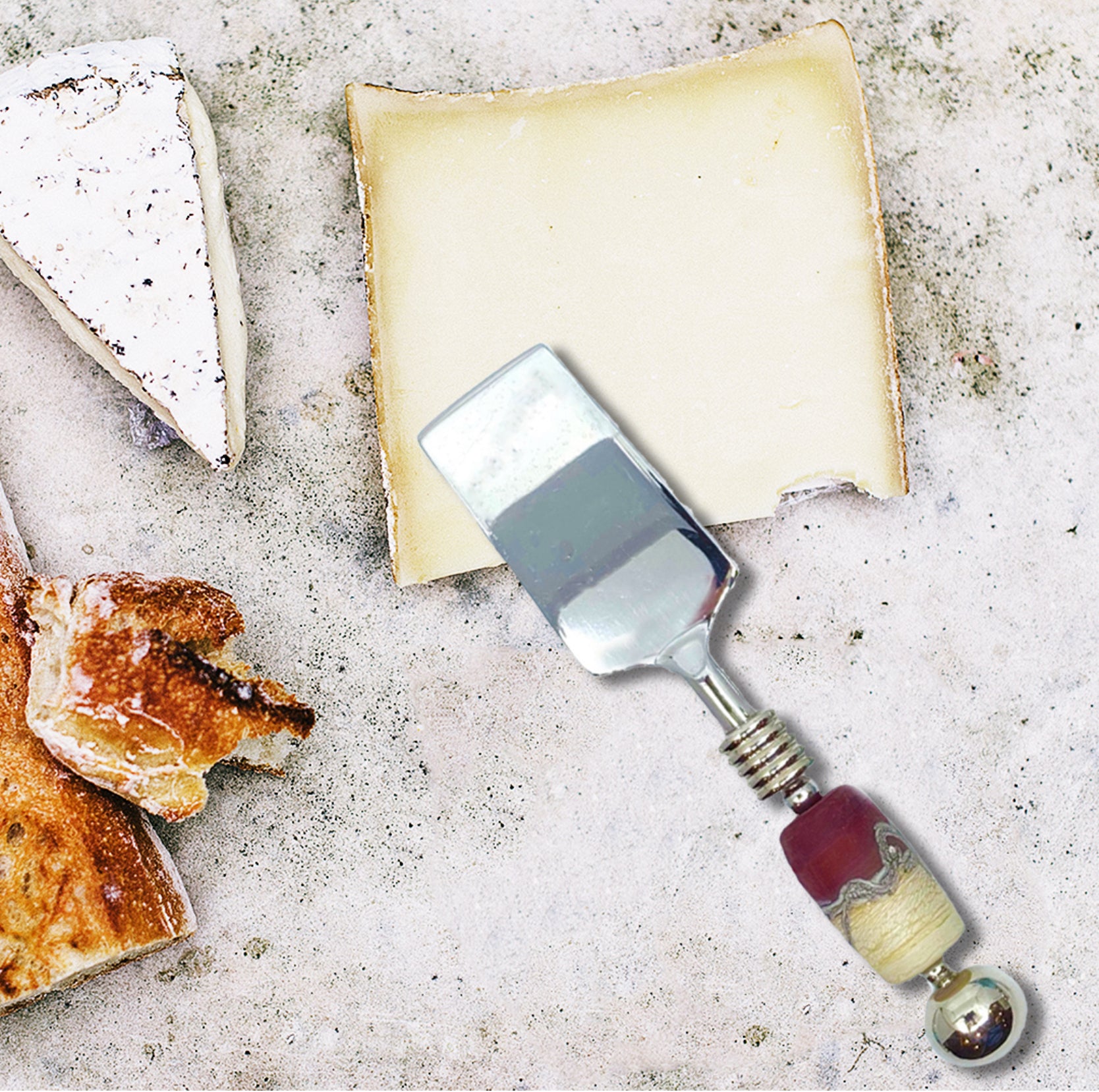 Cheese knife with decorative handle on a stone surface with cheese and bread.