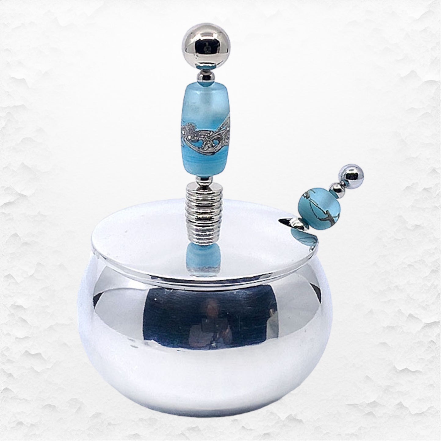 A handcrafted glass beaded sugar bowl with a stainless steel spoon, featuring a blue glass bead design and a silver-colored metal rim.