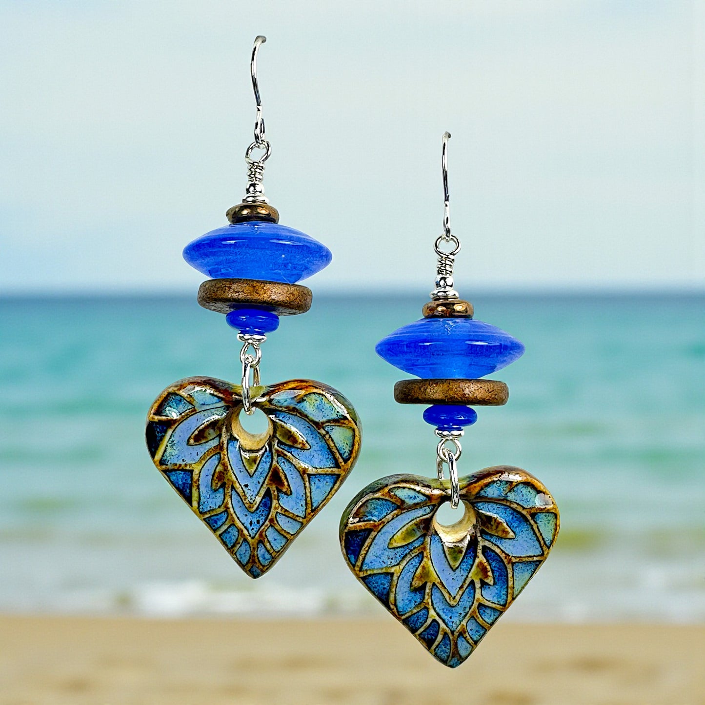 Heart-shaped earrings with blue and gold details against a beach background