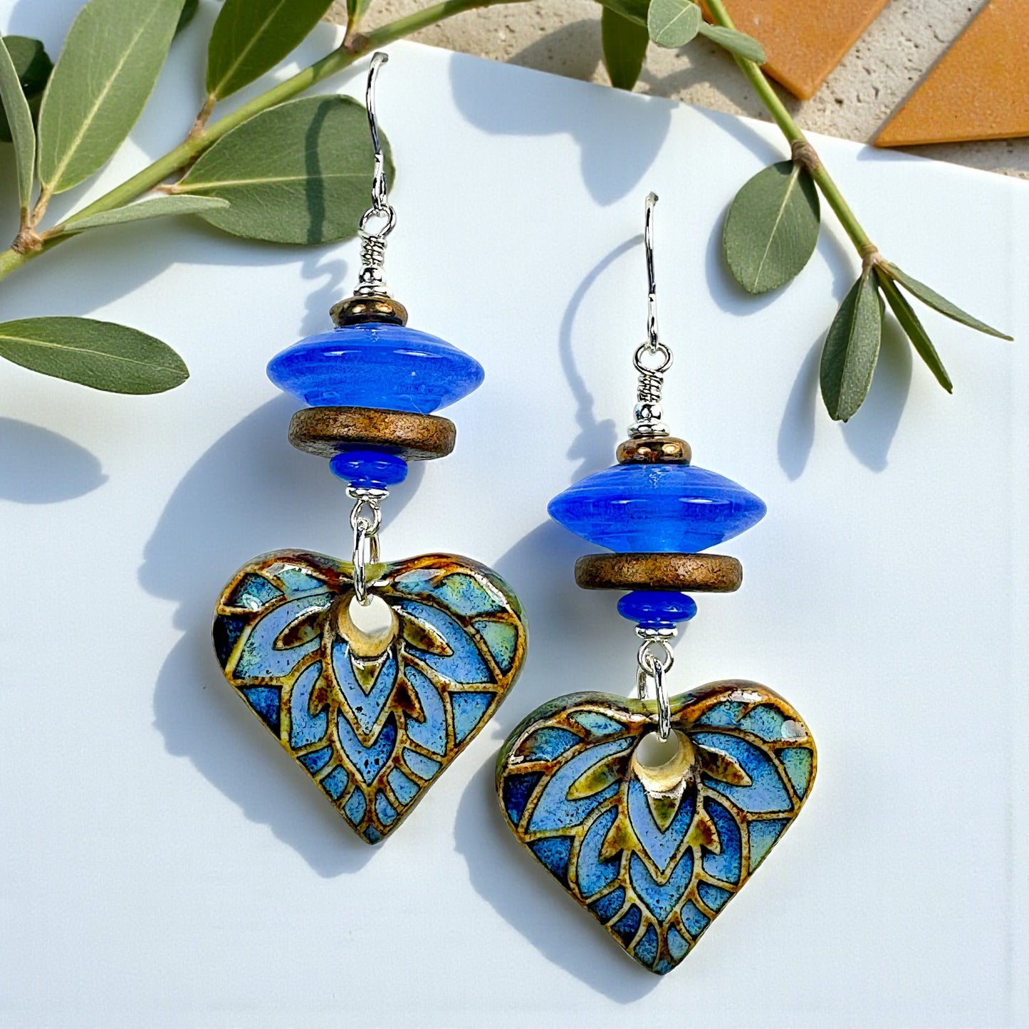 Heart-shaped earrings with blue and gold design on a white background with green leaves.