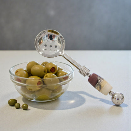 Silver ladle with decorative handle next to a glass bowl of green olives on a light surface.