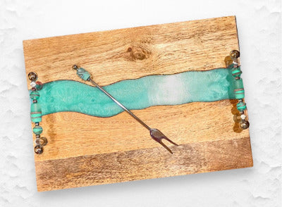 Handmade Glass Beaded Charcuterie Board.  

Each bead is individually shaped and arranged to create a beautiful accent that drapes effortlessly along your serving board. Perfect for parties, gifts, and elevating any table display with color, shine, and artisan charm.Wooden board with a decorative element featuring turquoise resin and gold accents on a white background