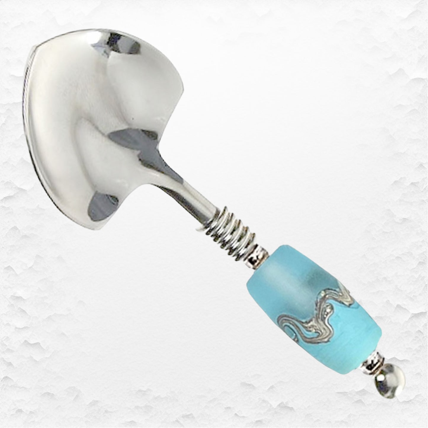 A stainless steel spoon with a small heart-shaped adorned with a turquoise handcrafted glass bead embellishment along the handle.