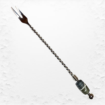 A long stainless steel pickle fork with a hand-torched glass bead design on the handle, featuring a brown handle and a green glass bead near the tip.