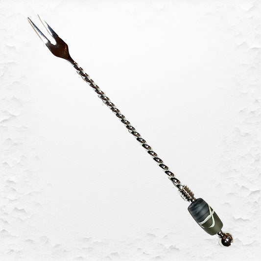A long stainless steel pickle fork with a hand-torched glass bead design on the handle, featuring a brown handle and a green glass bead near the tip.